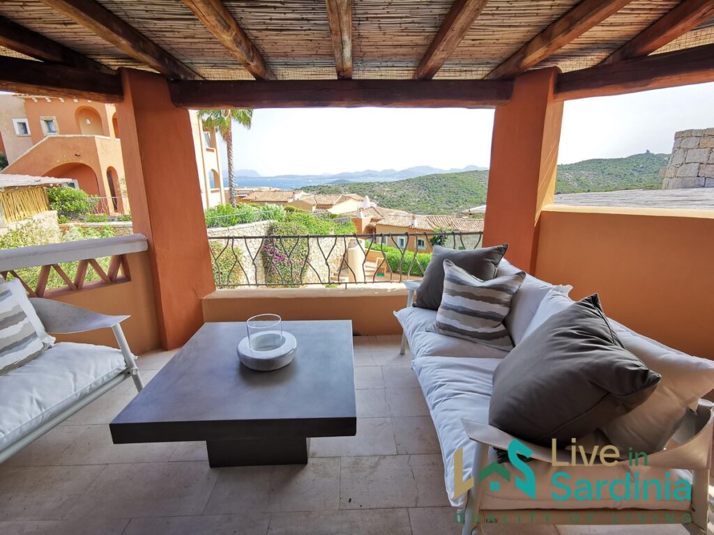 A terrace with table and sofa covered by a wooden roof framing a view on the coastline and natural surrounding