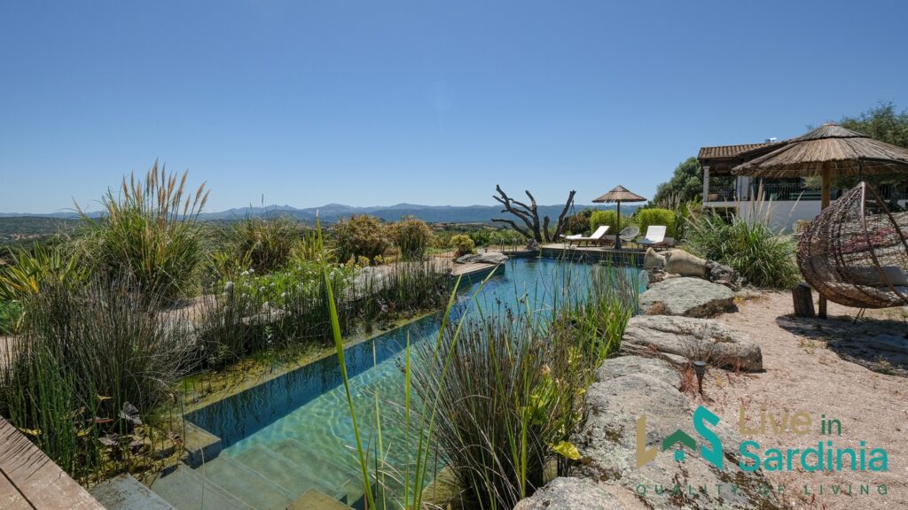 Bioclimatic pool surrounded by lush Mediterranean vegetation and granite rocks, with panoramic views of the Sardinian countryside and distant mountains, located at a countryside villa in Telti, Sardinia.