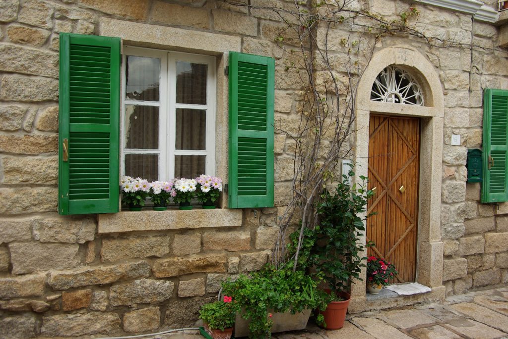 a green painted windows of a stone townhouse in sardinia with pot of flowers and wooden door with granite arch